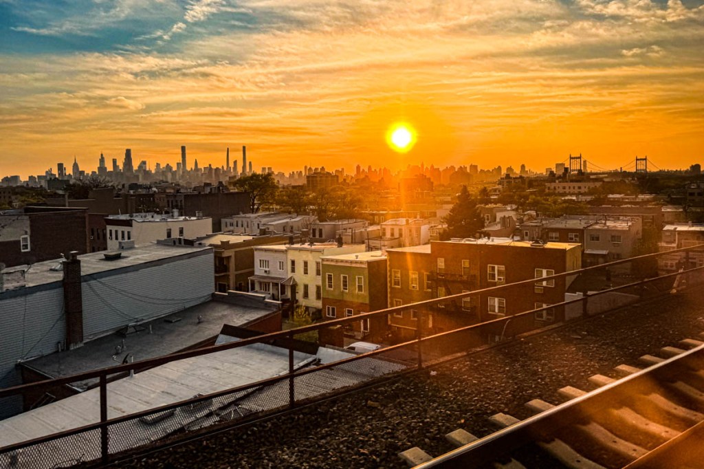 A vibrant sunset over the New York City skyline, with a golden sun setting on the horizon, surrounded by buildings and the silhouette of bridges.