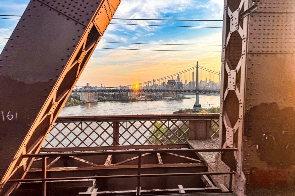 A view of the New York City skyline at sunset framed by the iron structure of a bridge, with water in the foreground reflecting the warm colors of the sky.