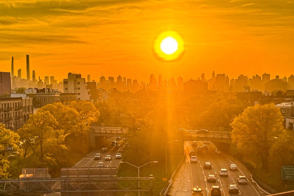 A vibrant sunset view over the New York City skyline, with the sun casting an orange glow and city buildings silhouetted against the colorful sky, accompanied by a tree-lined highway below.