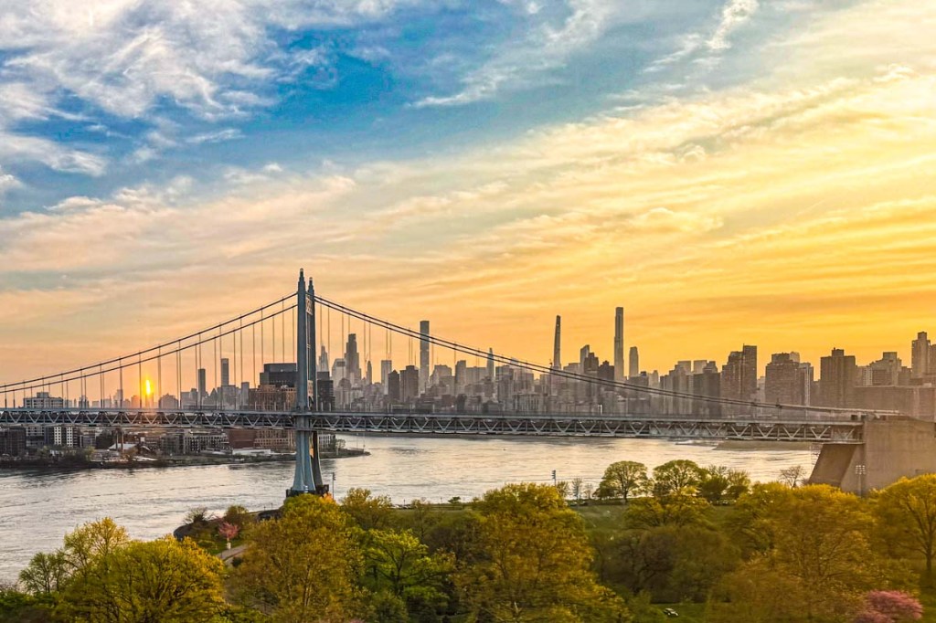 A scenic view of the New York City skyline at sunset, featuring a bridge crossing a river with skyscrapers in the background and vibrant orange hues in the sky.