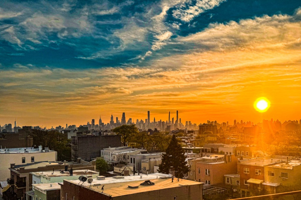 A stunning view of the New York City skyline at sunset, with vibrant orange and blue hues in the sky, casting a warm glow over the city. The image captures the silhouette of skyscrapers and rooftops in the foreground.